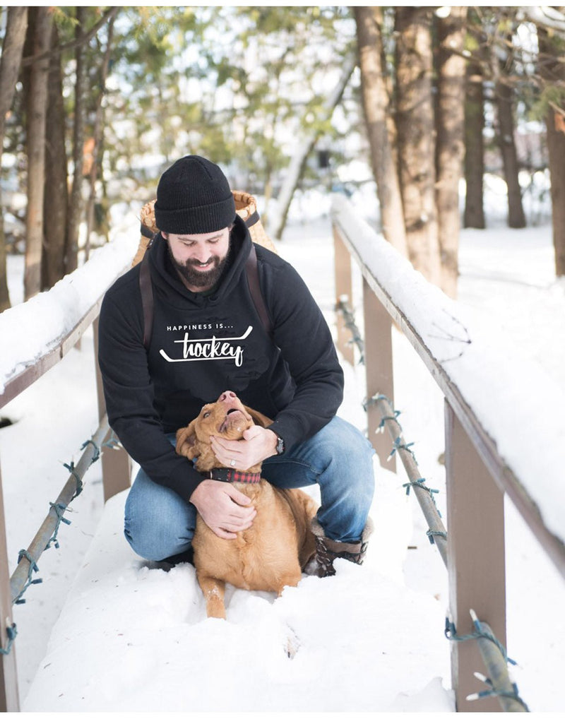 Man wearing blue jeans, a black toque, a backpack, and Happiness Is...Hockey Unisex Hoodie in jet black crouched down on a bridge in a snow covered forest petting a brown dog