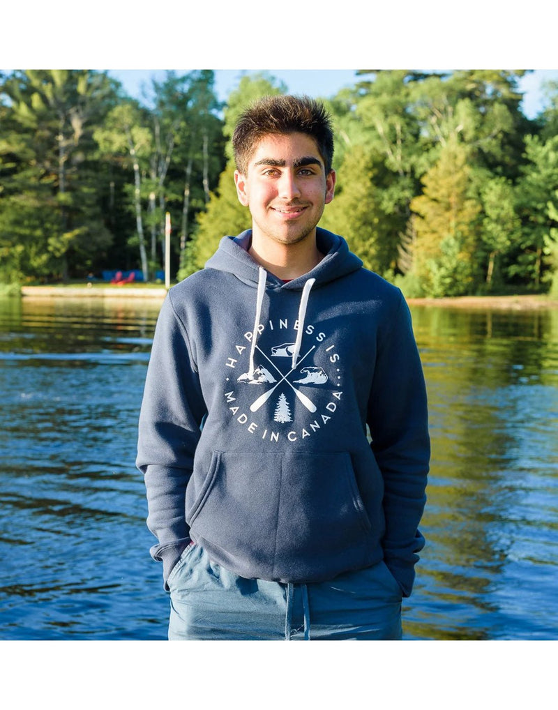 Man wearing blue shorts and Happiness Is...Unisex Crest Hoodie in navy, with his hands in his pockets and a lake and trees behind him