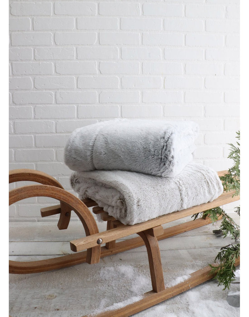 Lifestyle image of the Charcoal and Taupe frosted faux fur throws folded and stacked on top of a wooden sleigh with evergreen branches, in front of a white brick wall
