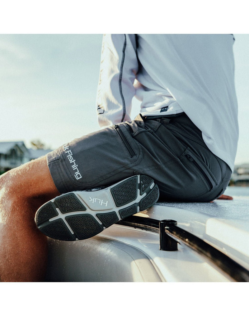 Lifestyle image of a man  sitting on a boat wearing a long sleeve top, Huk footwear,  and Huk Men's Next Level 7 inch Short Charcoal Grey.