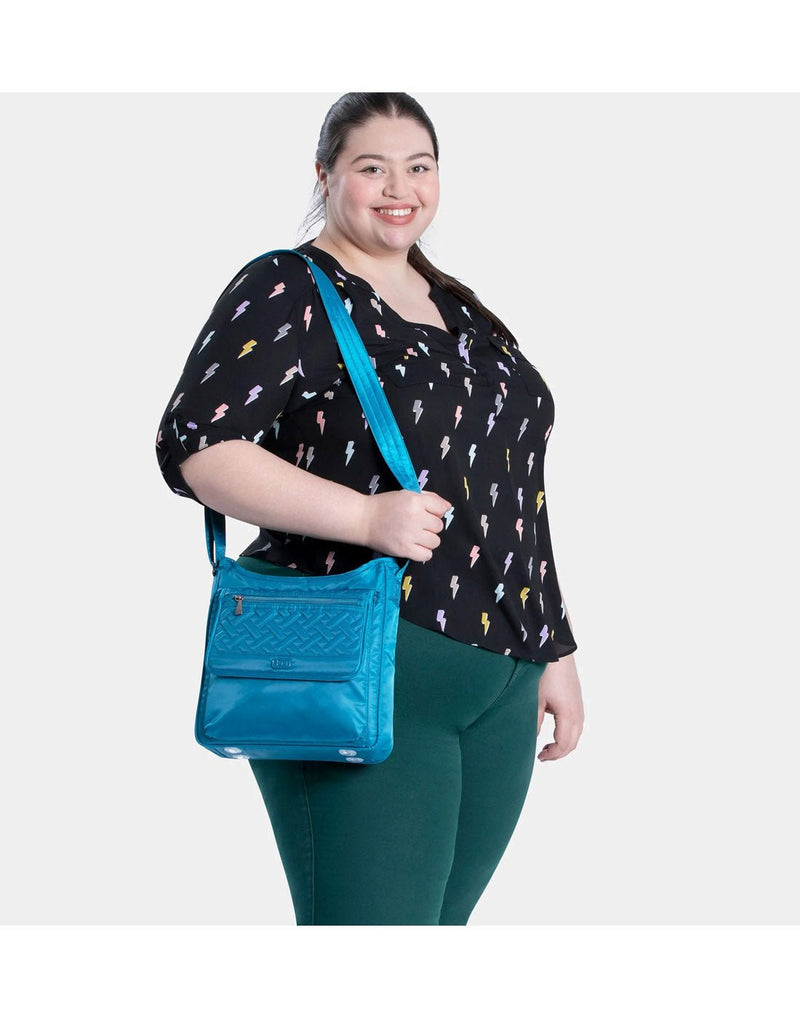 Woman wearing black blouse with white lightning bolts and green pants with Lug Hopscotch Crossbody Bag in ocean blue slung over one shoulder