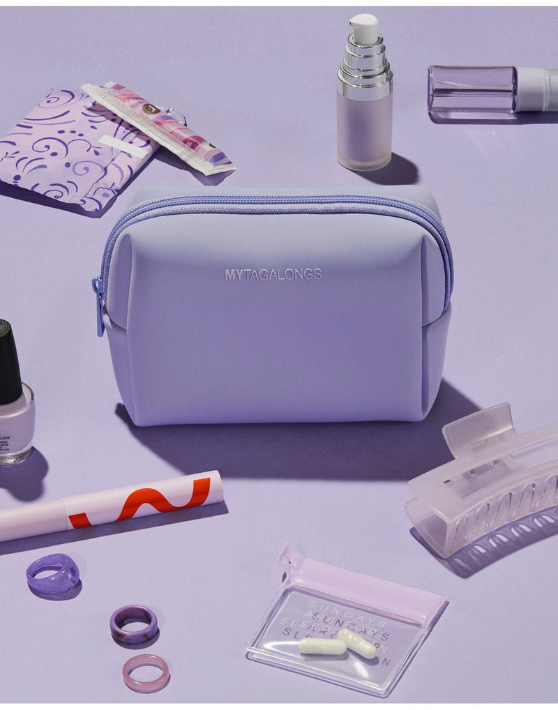 Purple product spread - light lilac cosmetic case on a lilac surface with hair clip, nail polish bottle and other small bottles, feminine products, rings and small zip pouch with pills all scattered around