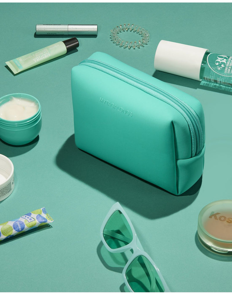Clover product spread - clover cosmetic case on a teal surface with make-up tubes and containers, sunglasses and hair tie scattered around