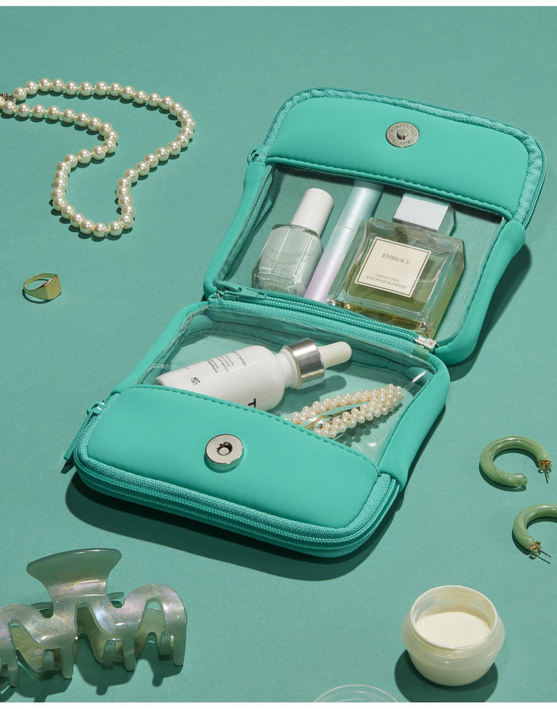 Clover product spread - double pouches, opened to clear sides inside, but still zipped together. There are other teal and green items scattered around like a pair of earrings, hair clip, gold ring, pearl necklace.  There is a small bottle of perfume, nail polish, dropper bottle, mascara and hair clip inside the pouches.