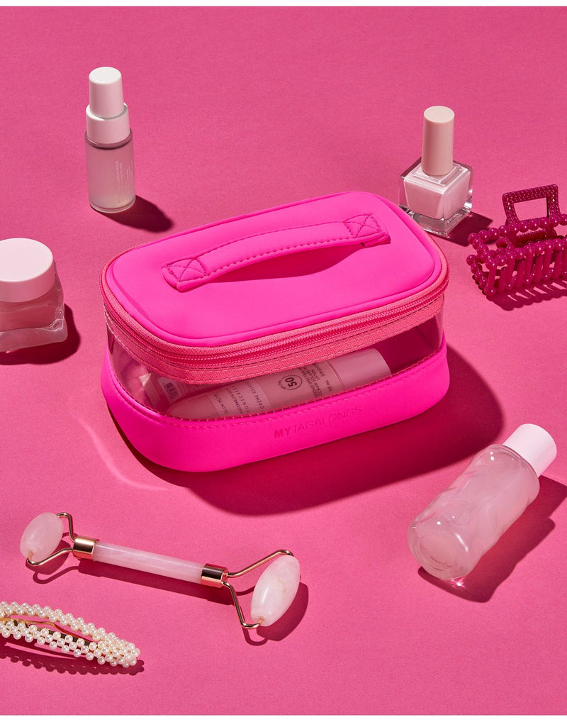 Pink product spread - hot pink train case on a hot pink surface with pink hair clip, several white and clear cosmetics bottles, a peal hair clip, and rose quartz roller spread around.