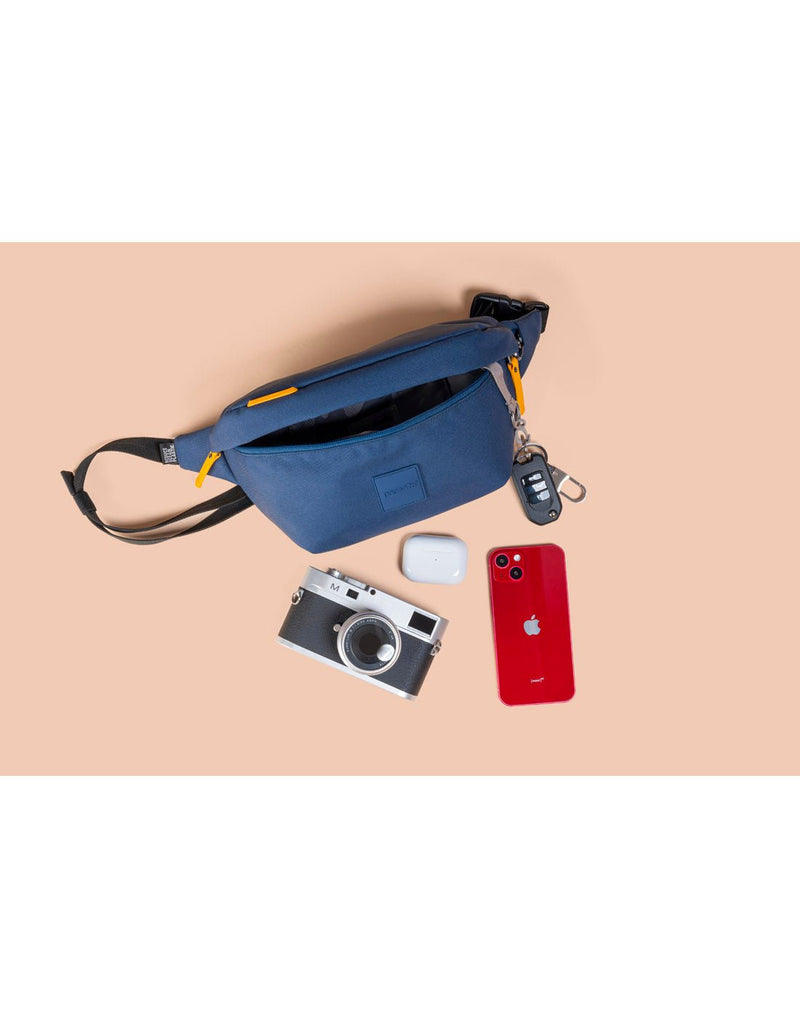 Product spread on peach background with Pacsafe®Go Anti-Theft Sling Pack in coastal blue, unzipped with key fob attached  and camera, ear pod case and red phone beside