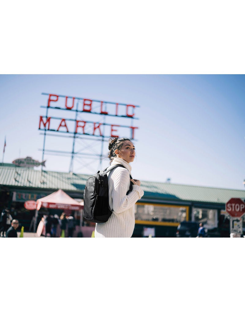 Lifestyle image of woman wearing a white turtleneck sweater and the black Pacsafe Metrosafe LS350 backpack at a public market with booths set up in the background