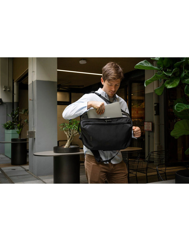 Lifestyle image of man wearing white shirt and tan pants standing outside an office cafe with the Pacsafe Metrosafe X Anti-Theft 16-Inch Commuter Backpack on one shoulder putting a laptop into bag in front of him