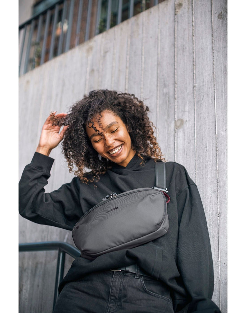 Lifestyle image of woman outside wearing black shirt and jeans with the Pacsafe Metrosafe X Anti-Theft Urban Sling in grey slung across her front