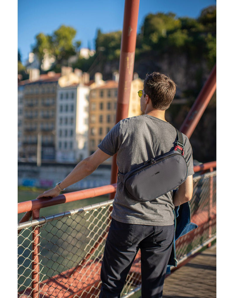 Lifestyle image of man wearing grey t-shirt and black jeans with the Pacsafe Metrosafe X Anti-Theft Urban Sling in slate, slung across his back as he stands looking out on a fenced bridge