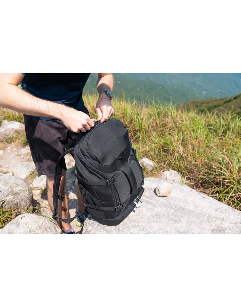 Lifestyle image of man standing outside on grass with the Pacsafe Venturesafe® EXP35 Anti-Theft Travel Backpack in black on a large rock, zipping up the bag
