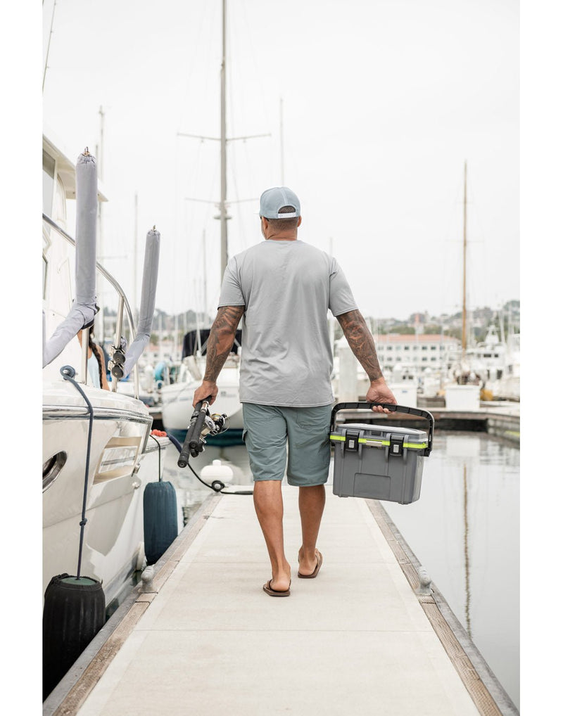 Lifestyle image of the back of a man with arm tatoos wearing a light grey t-shirt, sage green shorts, sandals and a baseball cap, walking down a dock at a marina.  There are boats around and one beside him.  He is holding a fishing rod in his left hand and the Pelican™ Elite 14qt Cooler in dark grey in his right hand.