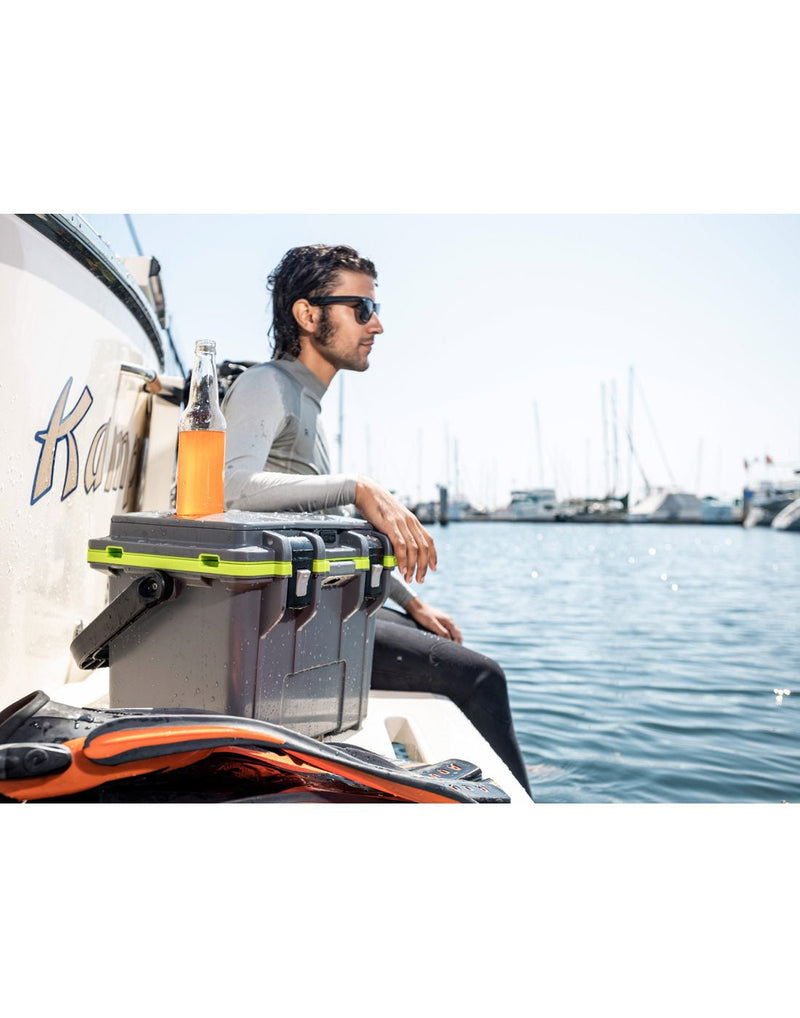 Lifestyle image of man sitting on the back of a boat wearing sunglasses, looking off to the right with his feet in the water.  The Pelican™ Elite 14qt Cooler in dark grey is beside him and he is resting his left arm on it.  There is a wetsuit beside the cooler and an opened orange drink bottle in a cupholder of the cooler.