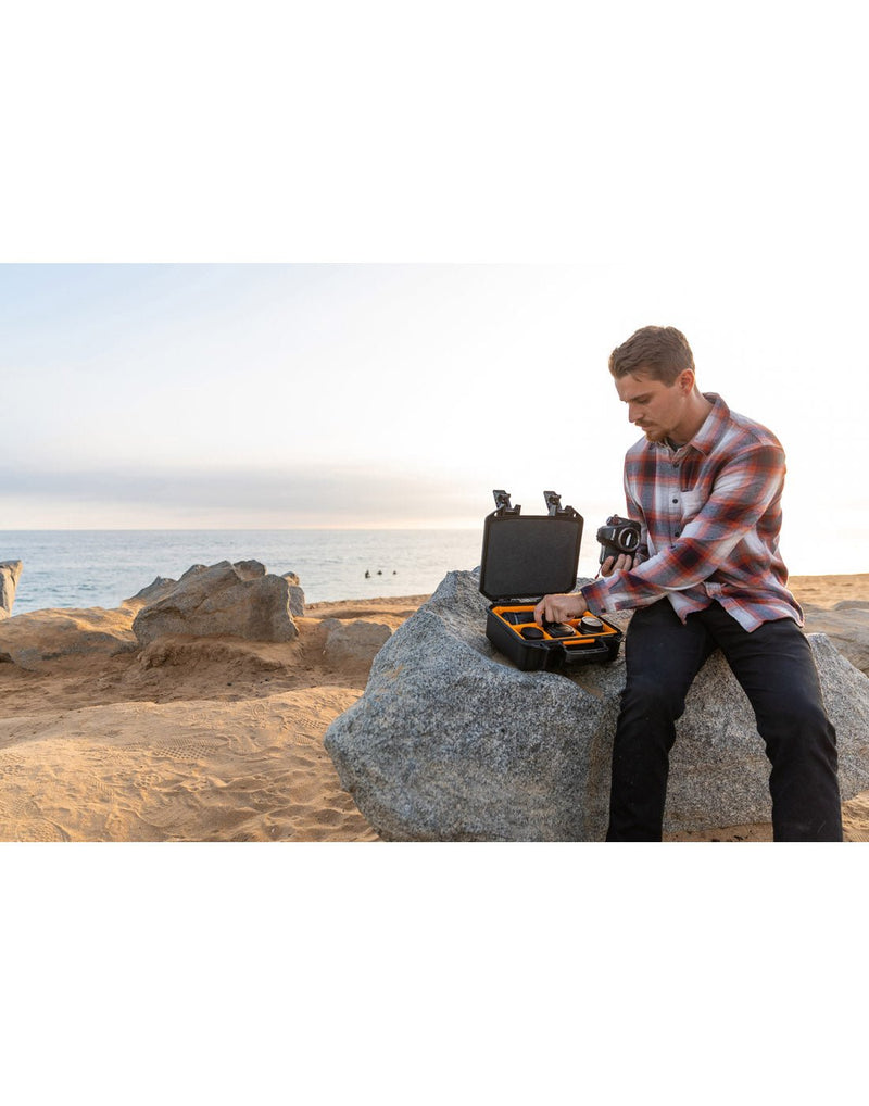 Lifestyle image of a man wearing black pants and a red, white and black plaid shirt, sitting on a large rock at a beach with the Pelican V200WD Vault Equipment Case open beside him on the rock as he is holding a camera and taking a camera lense out of the case