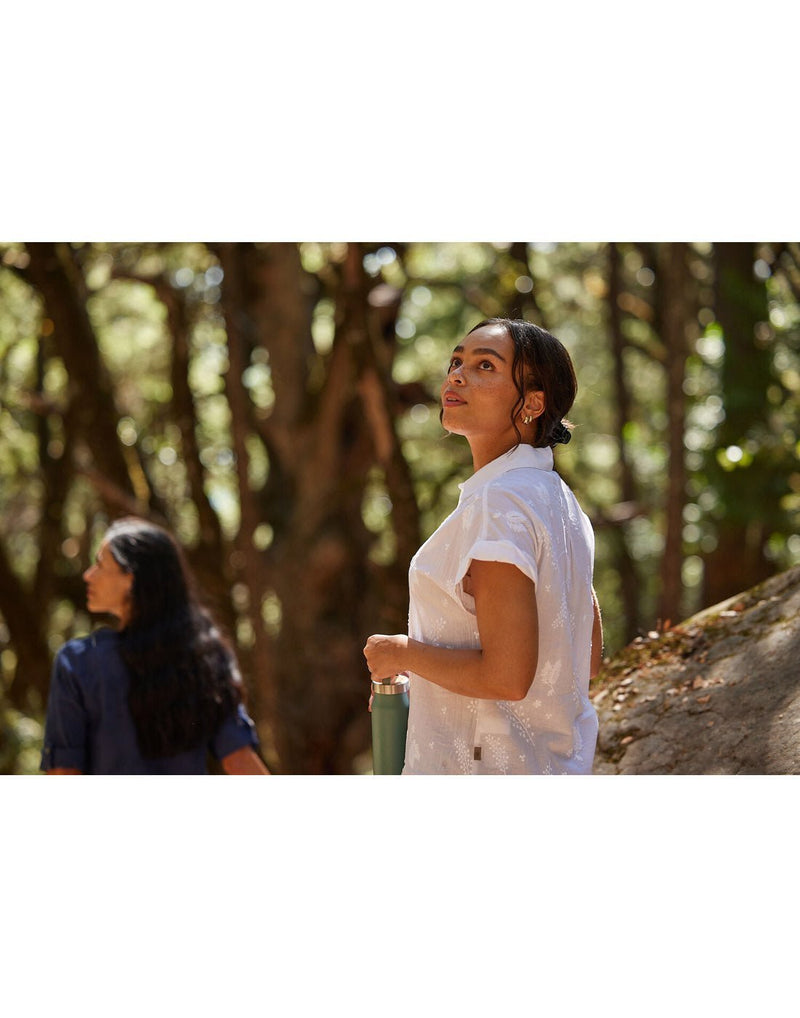Lifestyle image of woman walking in forest wearing Royal Robbins Women's Oasis Short Sleeve in white, holding water bottle and looking up