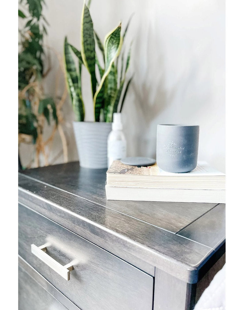 Lifestyle image of dark grey candle on top of two stacked books on a dark wood beside table with a potted plant beside